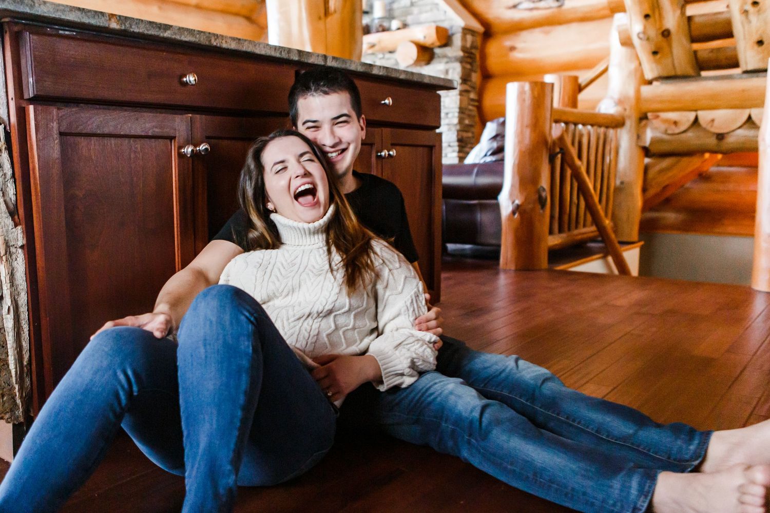 Two people wearing cozy sweaters laugh together while sitting on wooden floor in rustic log cabin interior.