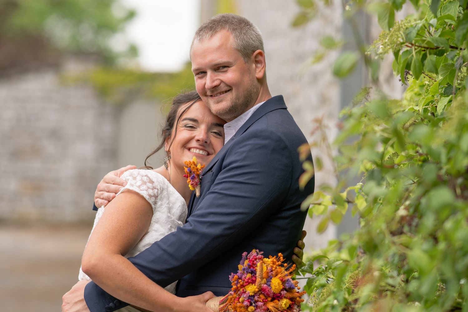 A joyful wedding couple embraces outdoors with autumn-colored flowers and green foliage in the background.