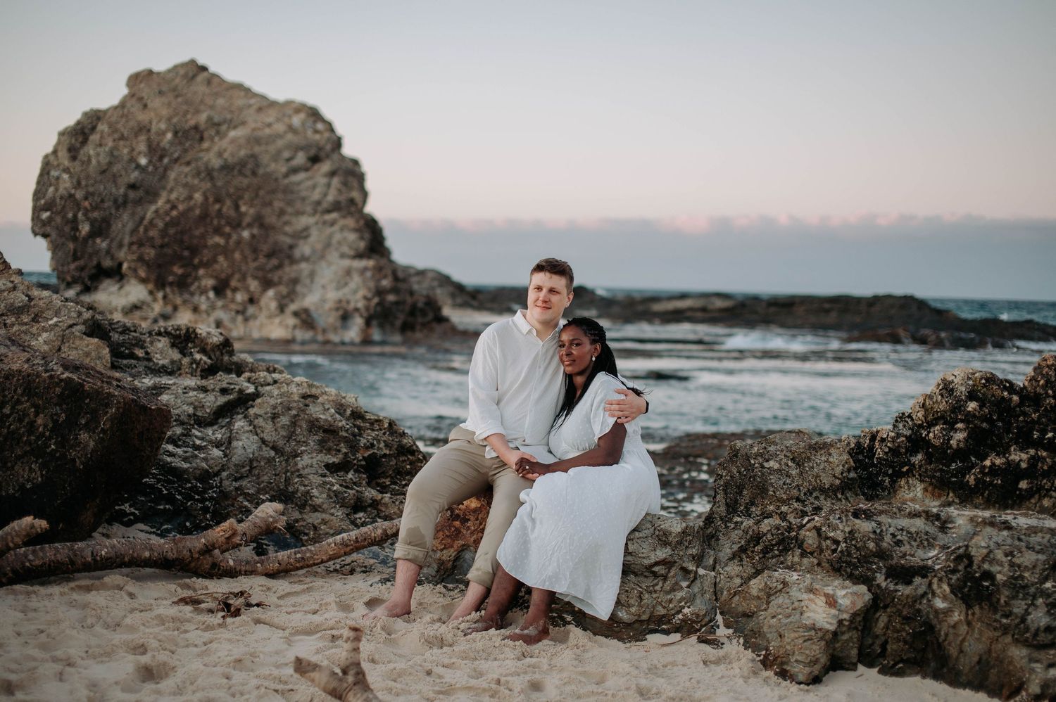 A couple sits together on coastal rocks at sunset overlooking the ocean and horizon.