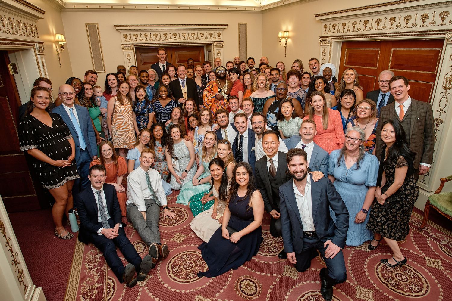 Large group of people posing for a panoramic photo inside an elegant room with ornate carpeting and wood paneling.