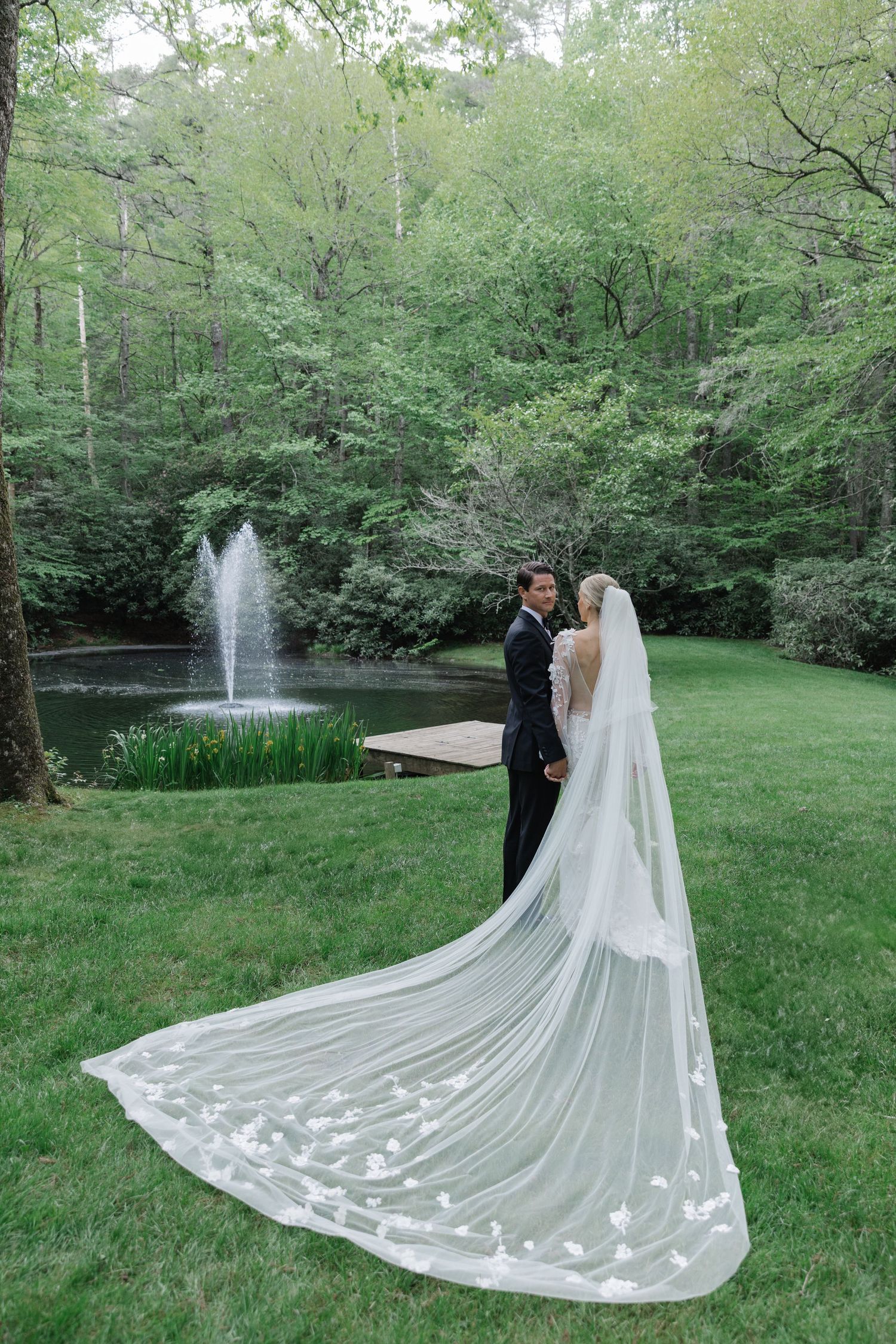 A couple in wedding attire stands in a lush garden setting with a flowing cathedral veil trailing behind them on the grass. Old Edwards Inn