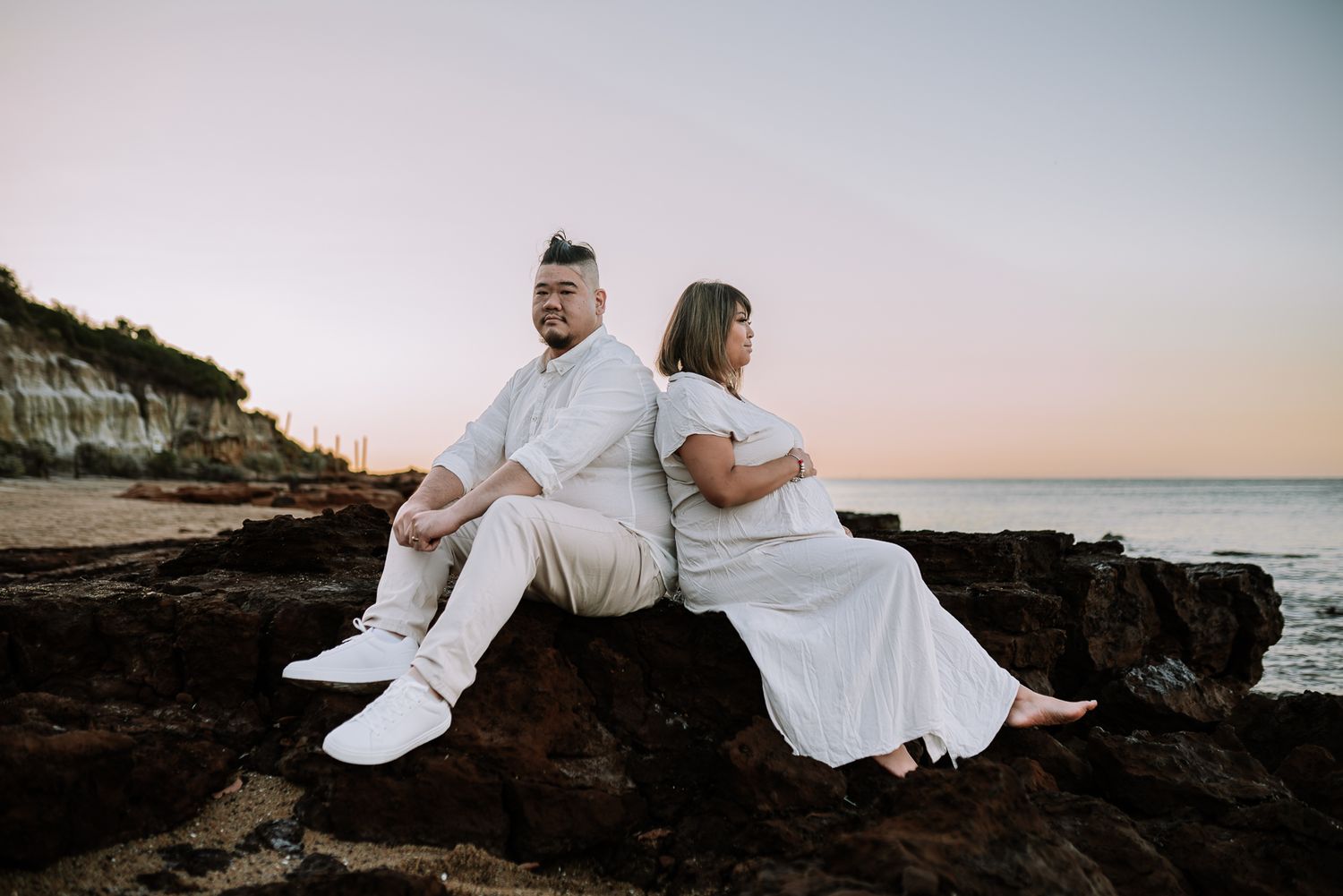 A couple in white outfits sits together at sunrise on a beach with ocean waves in the background taken by a Melbourne maternity photographer