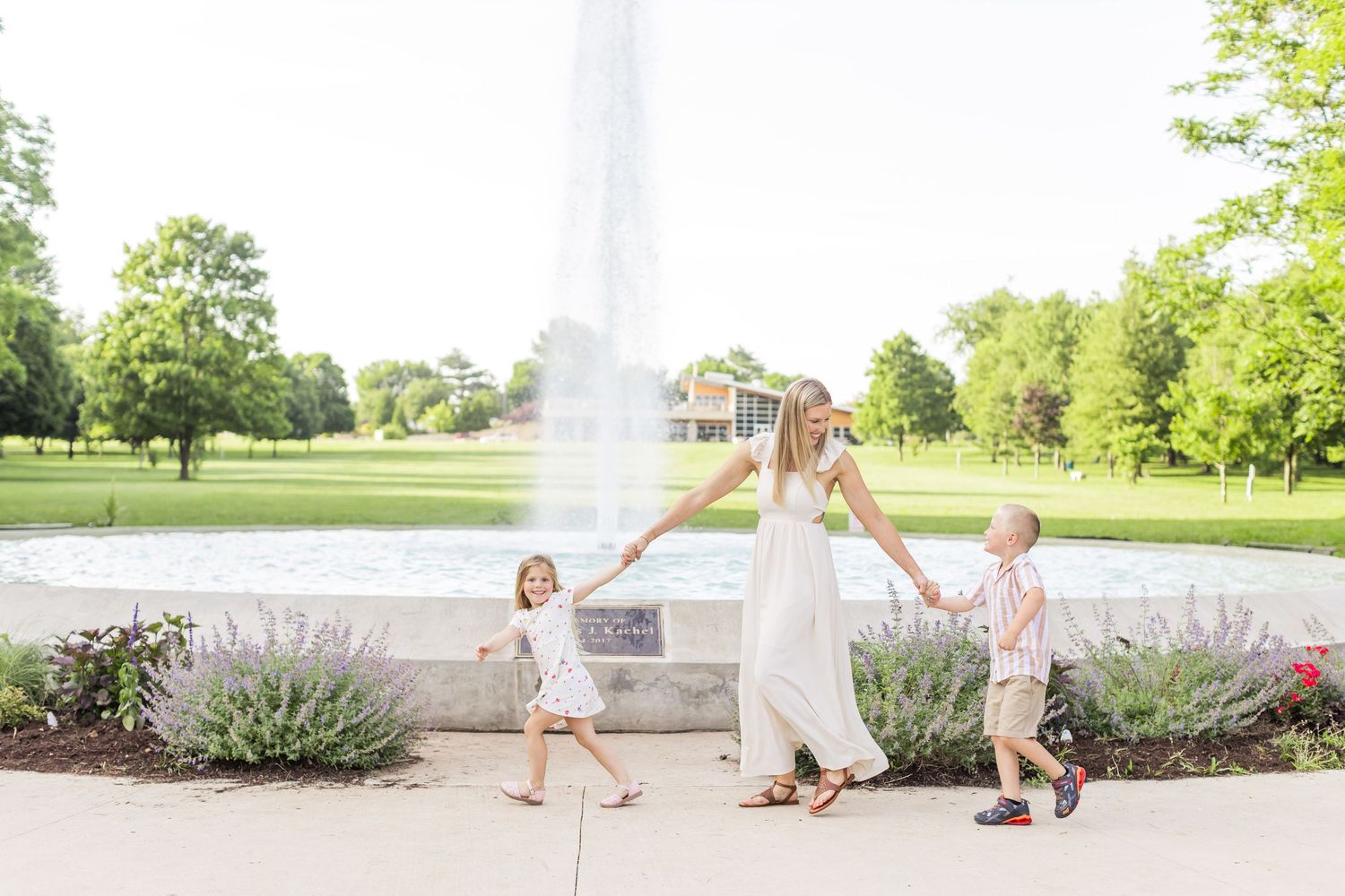 A family walks together on a sunny day in a park with a fountain and lavender plants in the foreground.