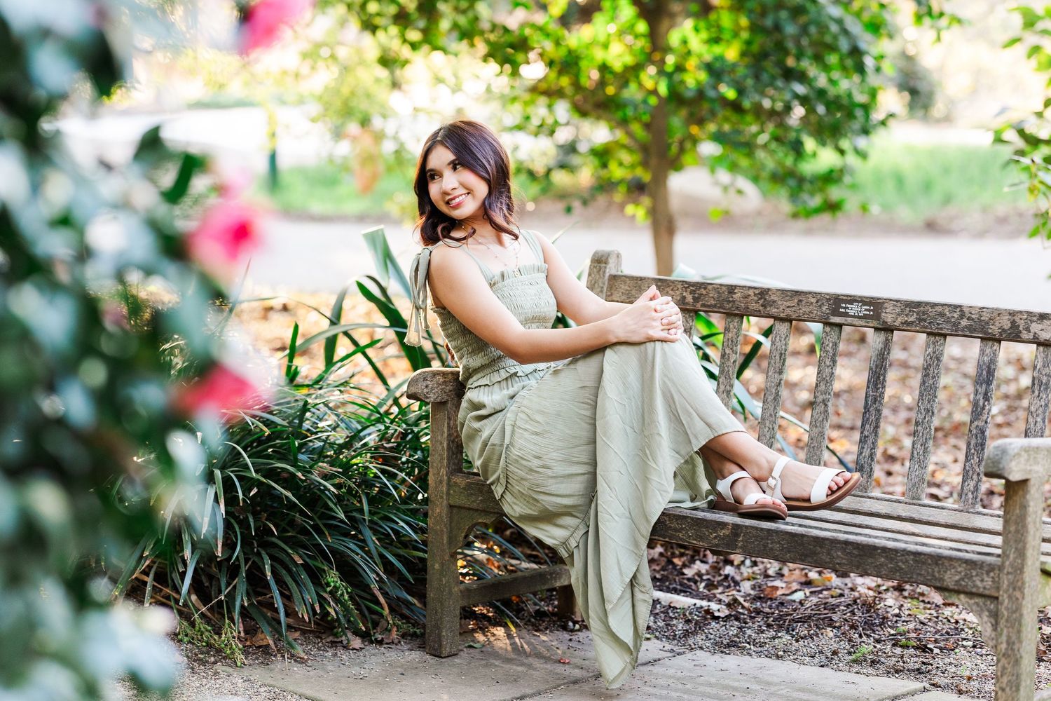 A woman in a beige dress relaxes on a wooden garden bench surrounded by pink flowers and greenery.