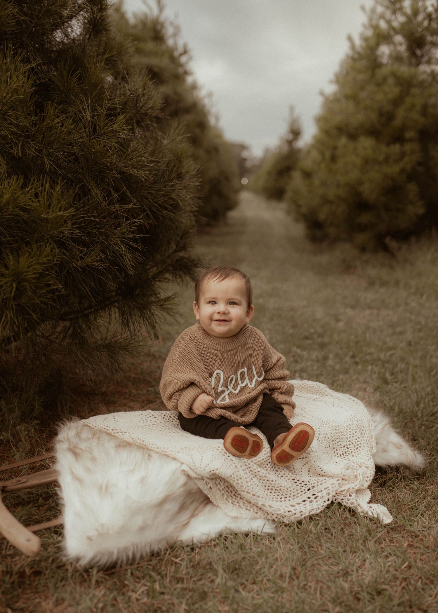 A baby in a brown sweater sits on a white blanket in a rustic outdoor setting with evergreen trees.