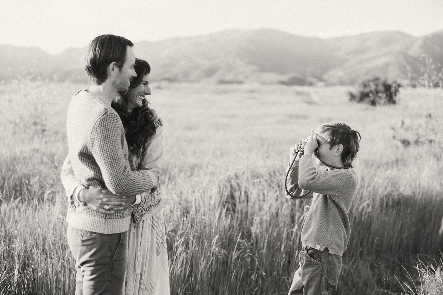 A candid black and white photo of a family moment in a grassy field with mountains in the background.