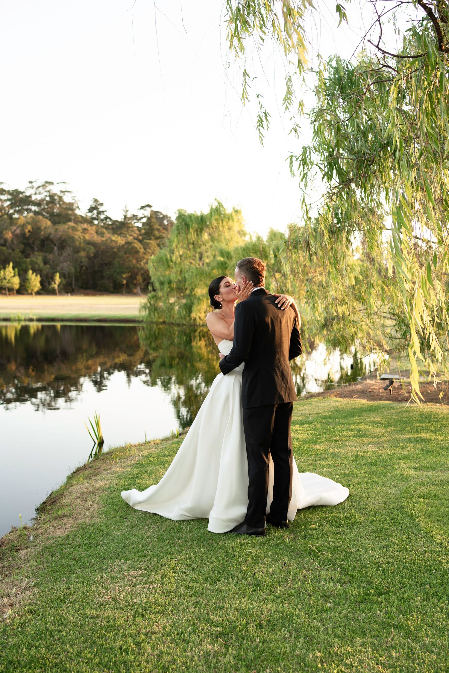 A couple in wedding attire embraces by a serene lake at sunset, with drooping willow branches framing the romantic scene.