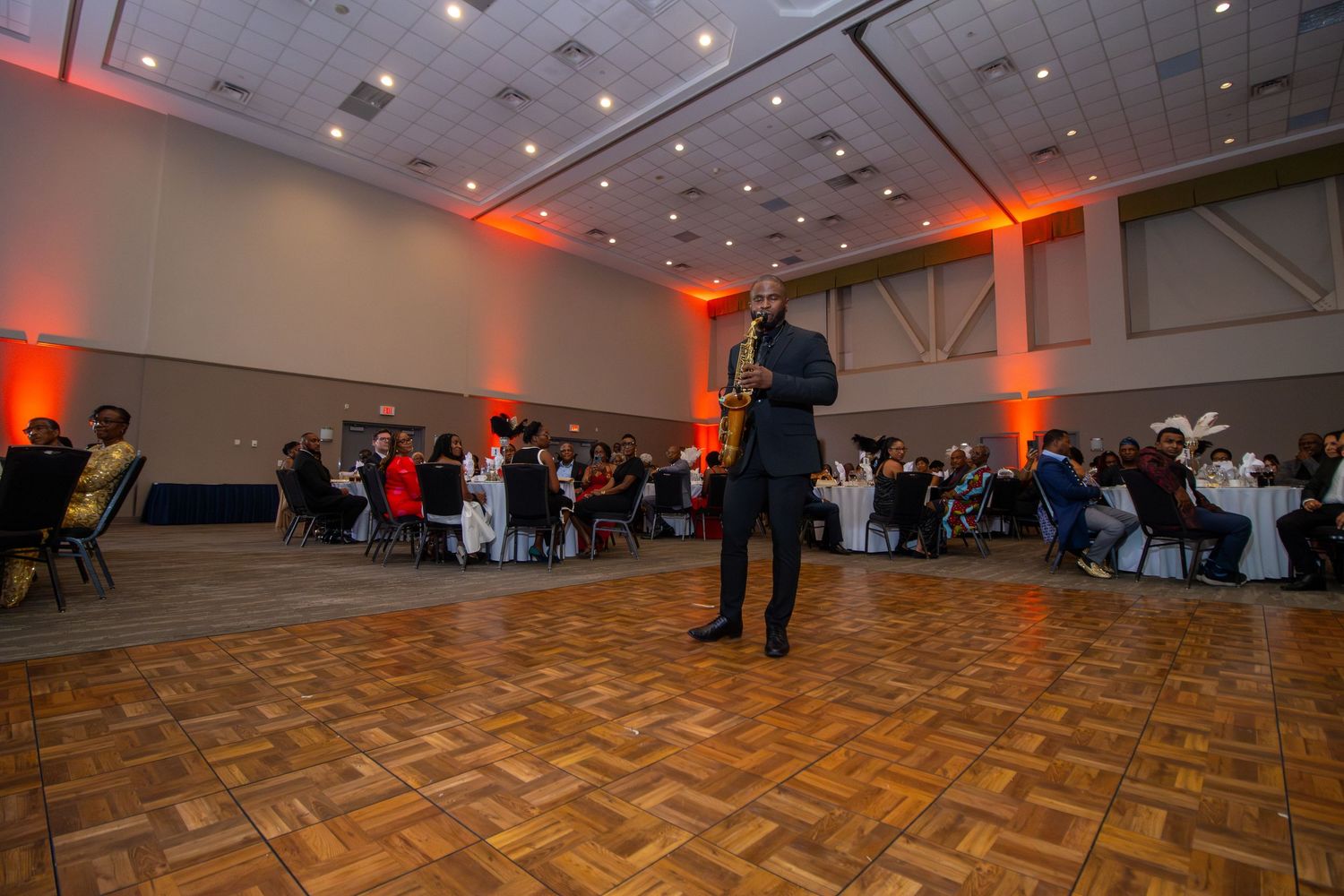 A speaker addresses attendees in a banquet hall with orange uplighting and parquet flooring during an evening event.