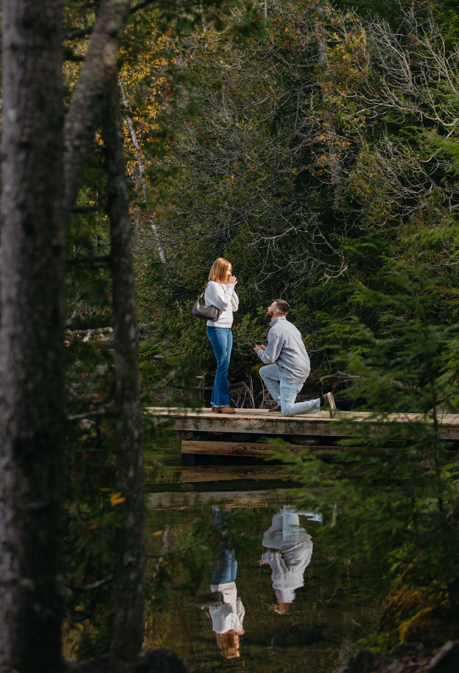 A romantic proposal scene unfolds on a wooden dock overlooking a tranquil pond surrounded by autumn trees.