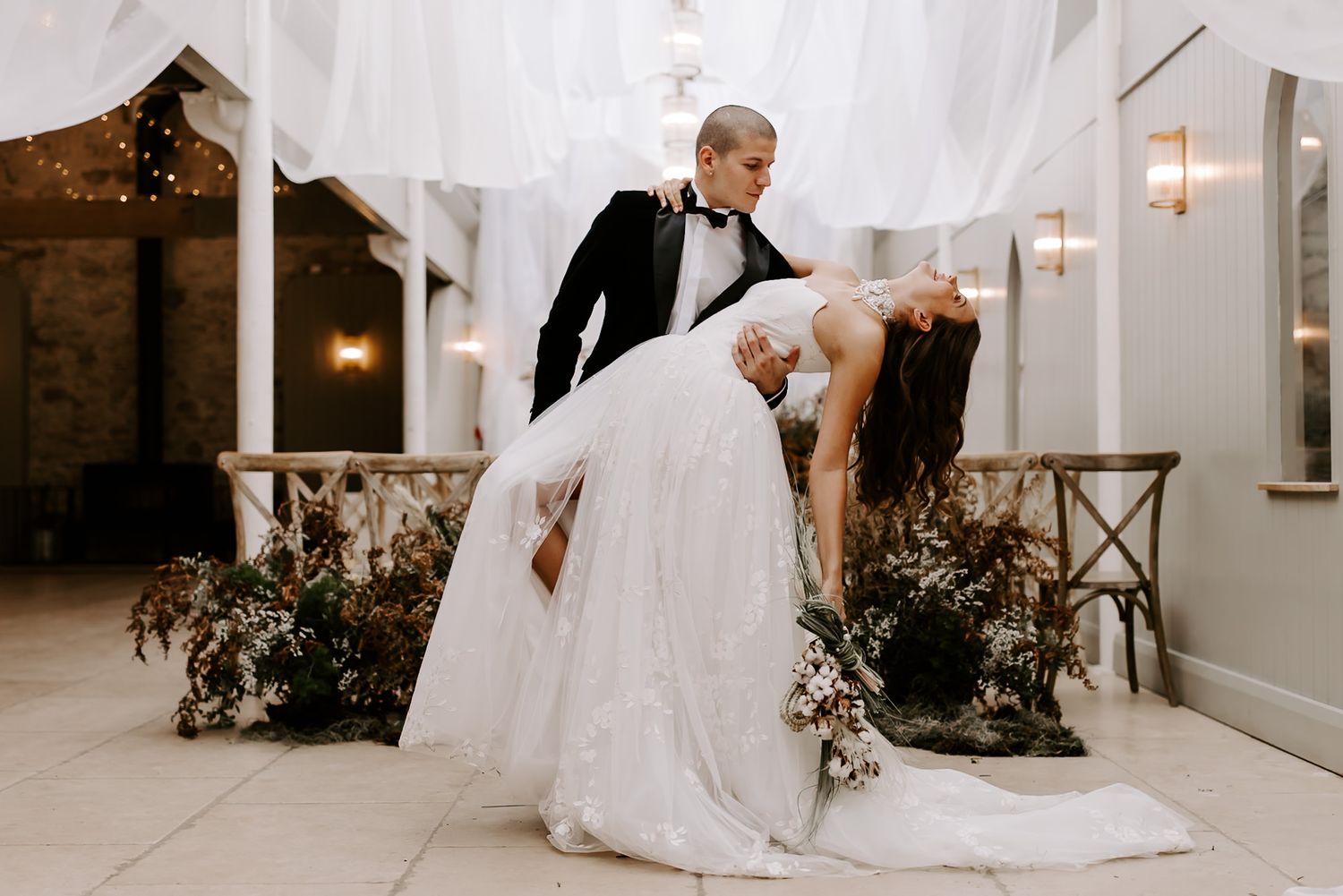 A romantic wedding couple performs an elegant dip dance move on a white patio with string lights and greenery.