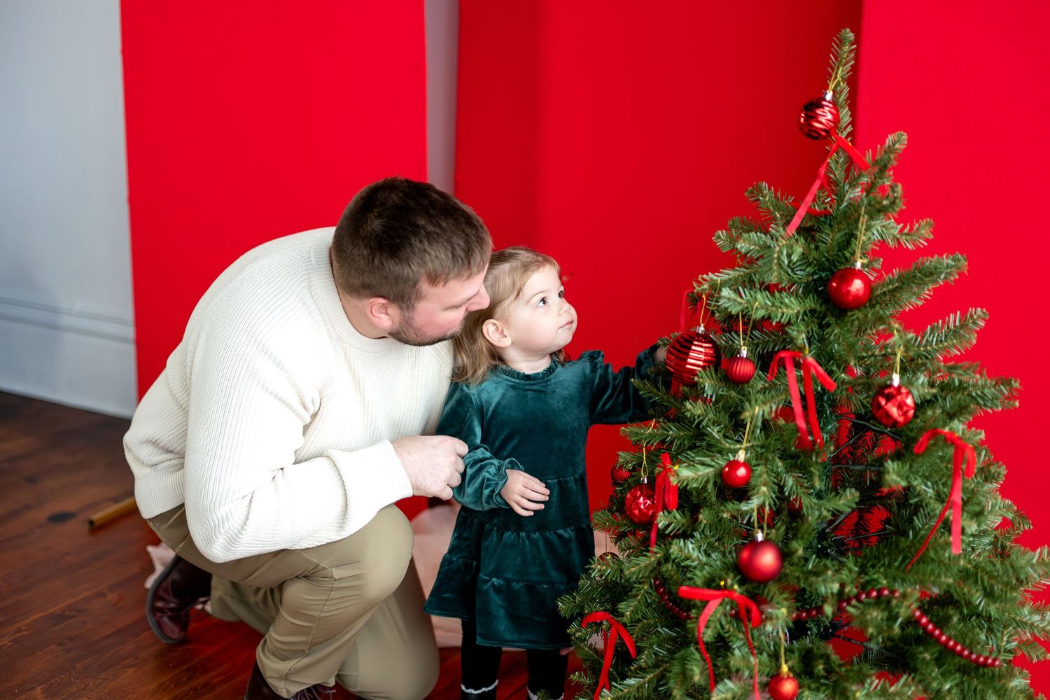 Father crouches beside toddler girl in green dress decorating Christmas tree with red ornaments.