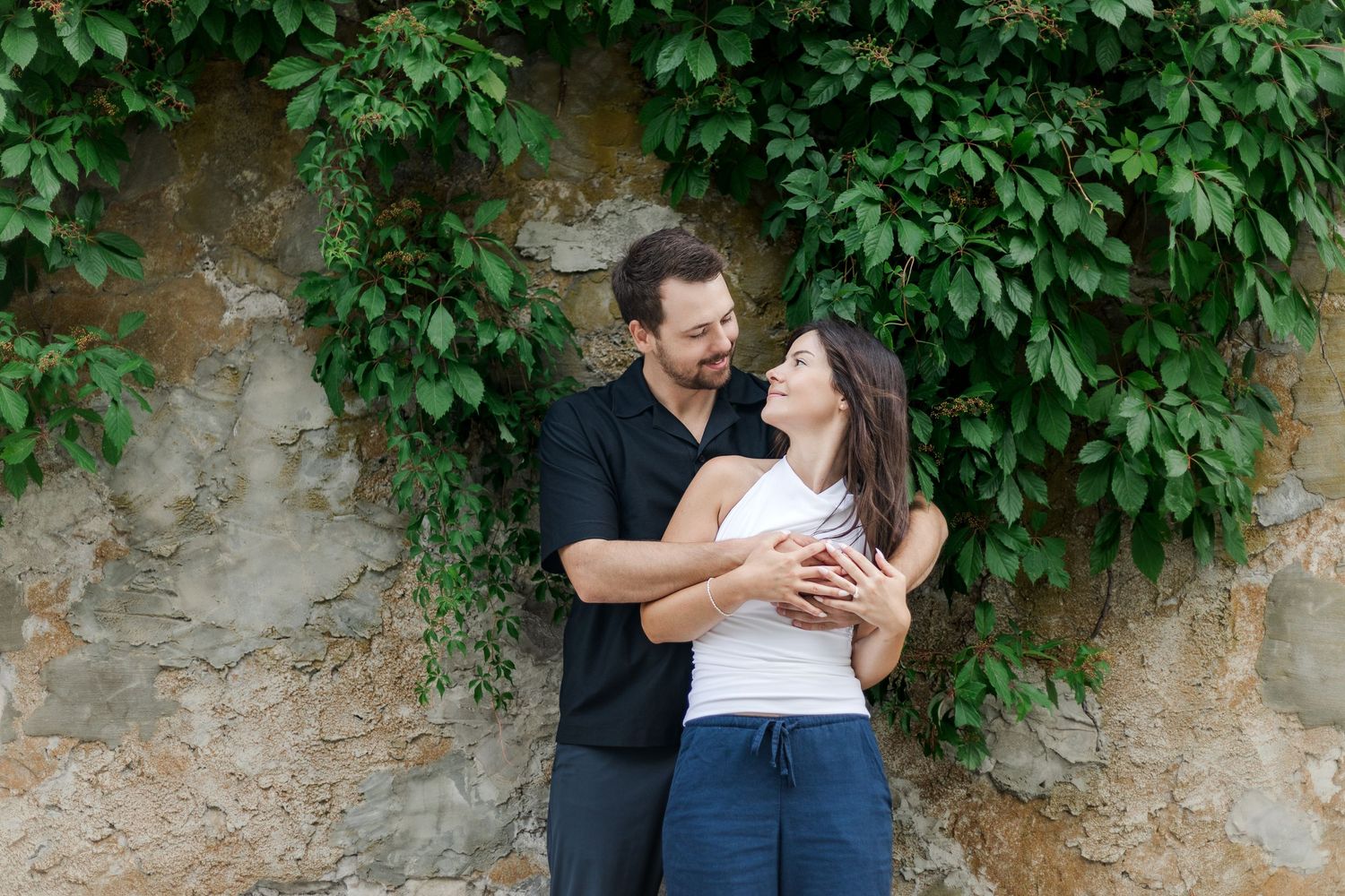 Romantic couple embracing against an ivy-covered stone wall in natural lighting.