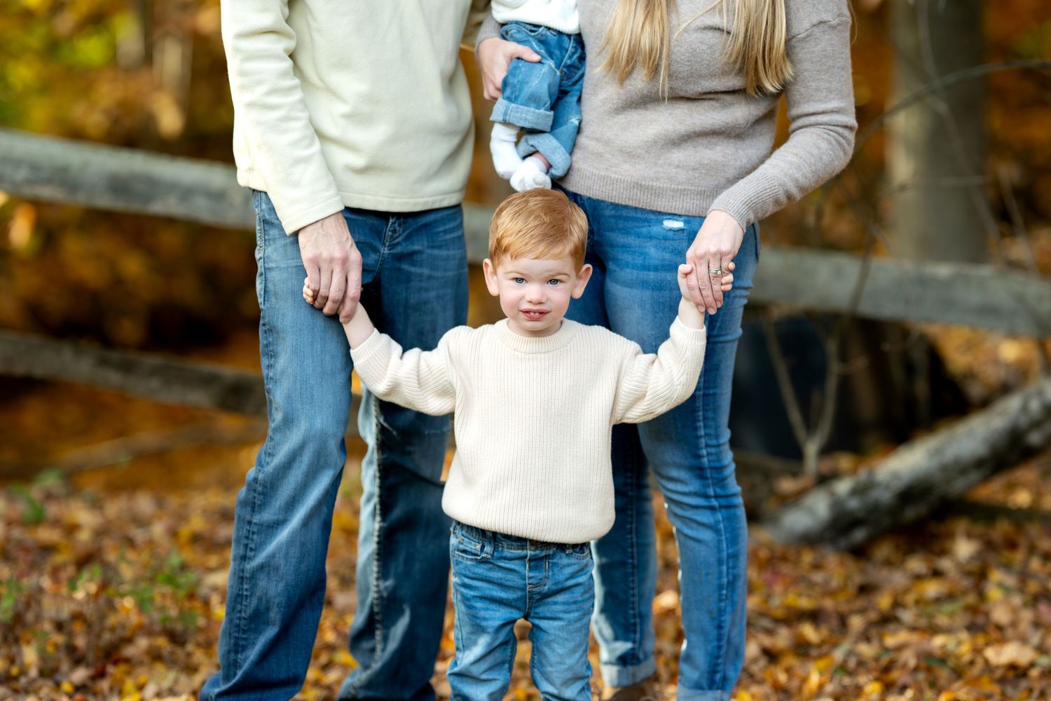 Toddler in white sweater holding parents hands in an autumn forest setting.