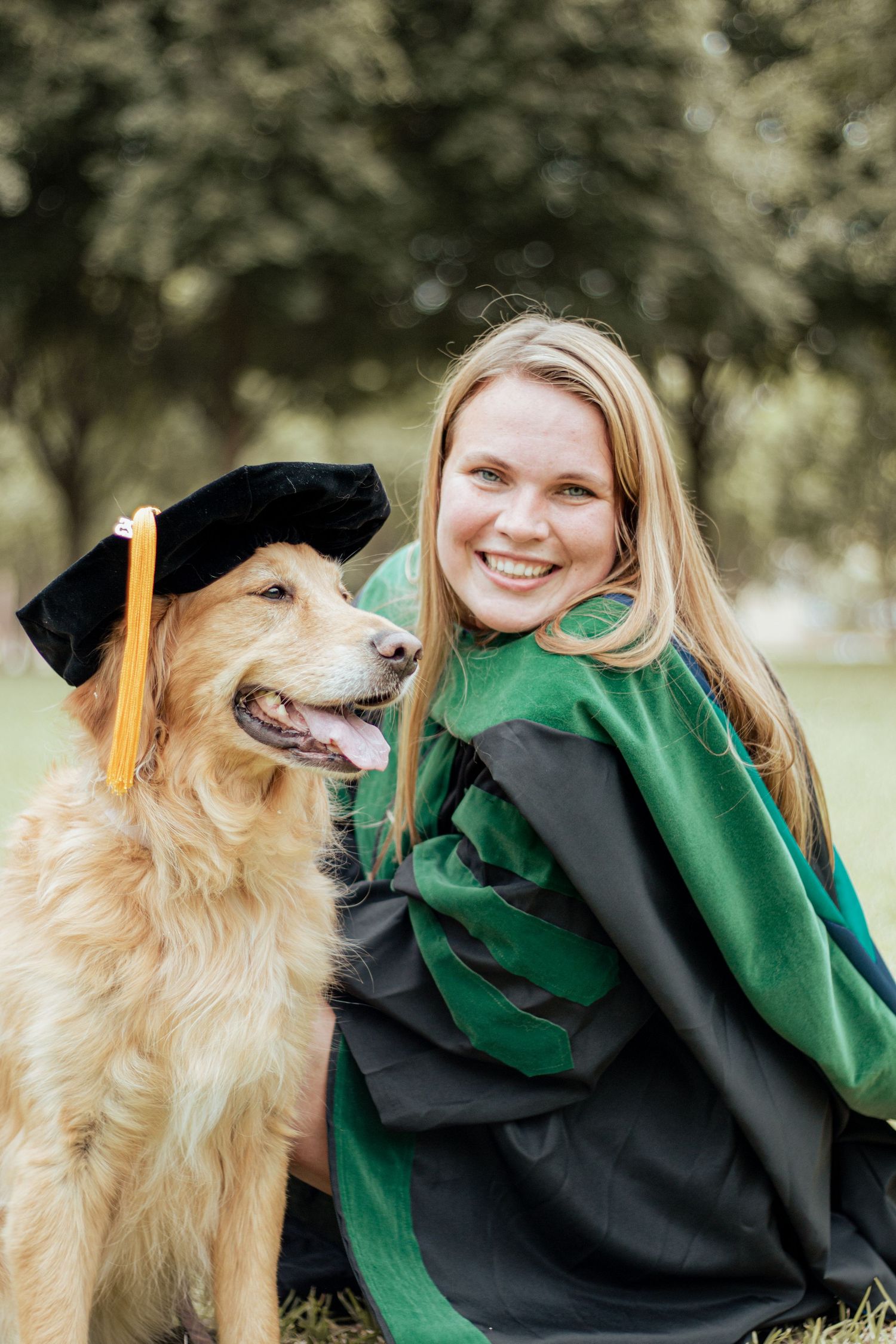 A golden retriever wearing a graduation cap poses with a graduate in green and black academic regalia outdoors.