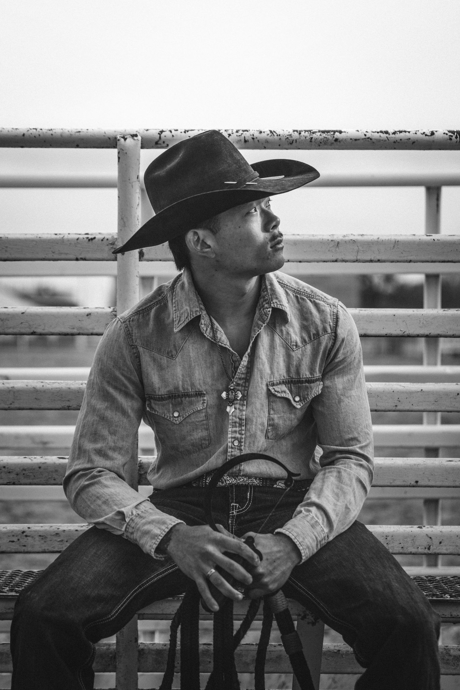 Black and white photo showing a cowboy in denim and western hat sitting pensively on metal rodeo gates.