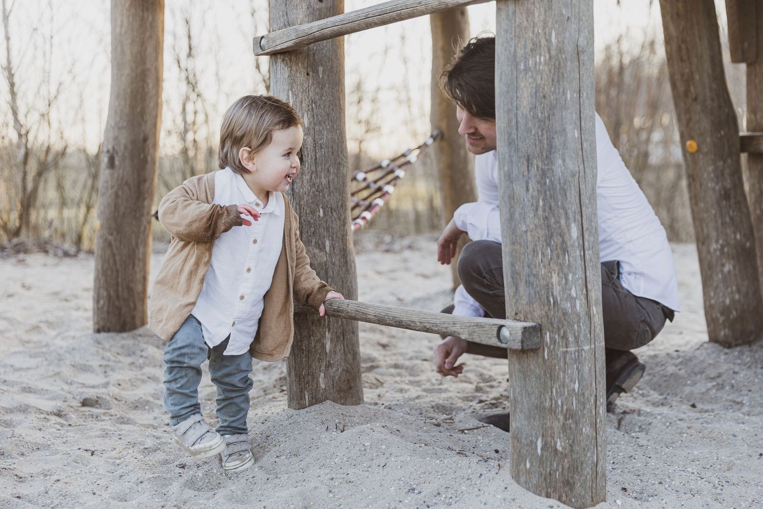 Two people enjoy a playful moment between wooden posts on a sandy ground in a rustic outdoor setting.