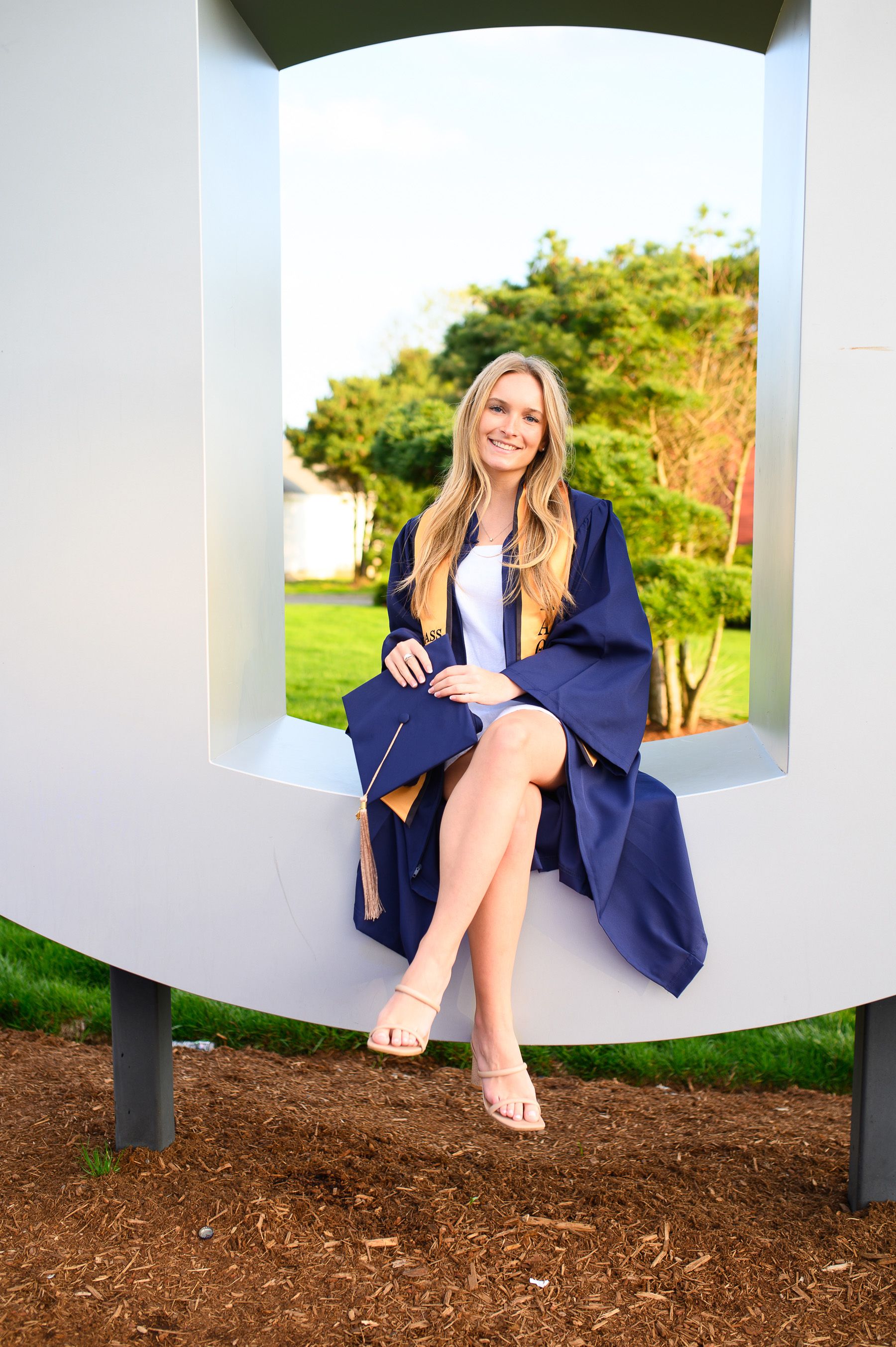 Graduate sits on modern outdoor bench in navy blue graduation gown and nude heels on sunny day.