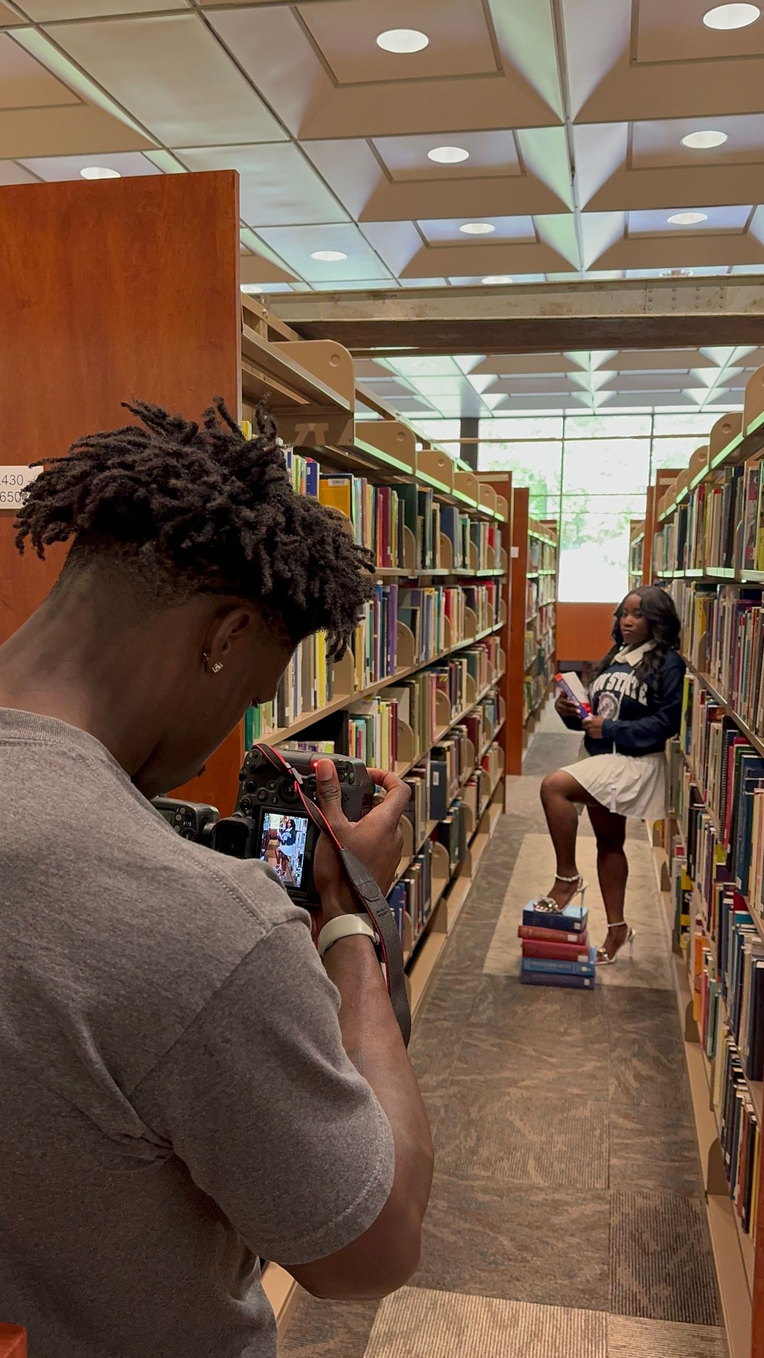 Students taking photos between bookshelves in a modern library with wooden shelving and fluorescent lighting.