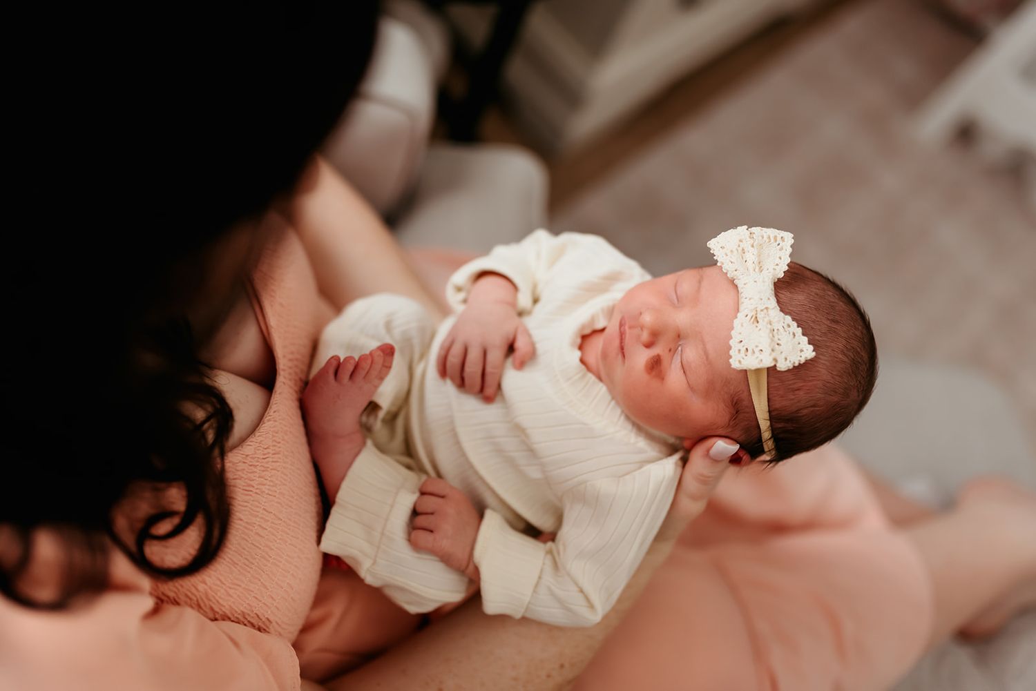 A sleeping newborn baby wearing a white outfit with a bow headband is held in her mother's arms.
