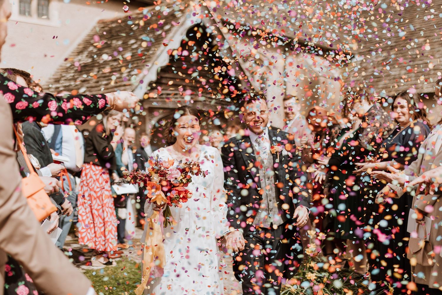 Happy newlyweds walk through shower of confetti while guests celebrate during outdoor wedding ceremony.