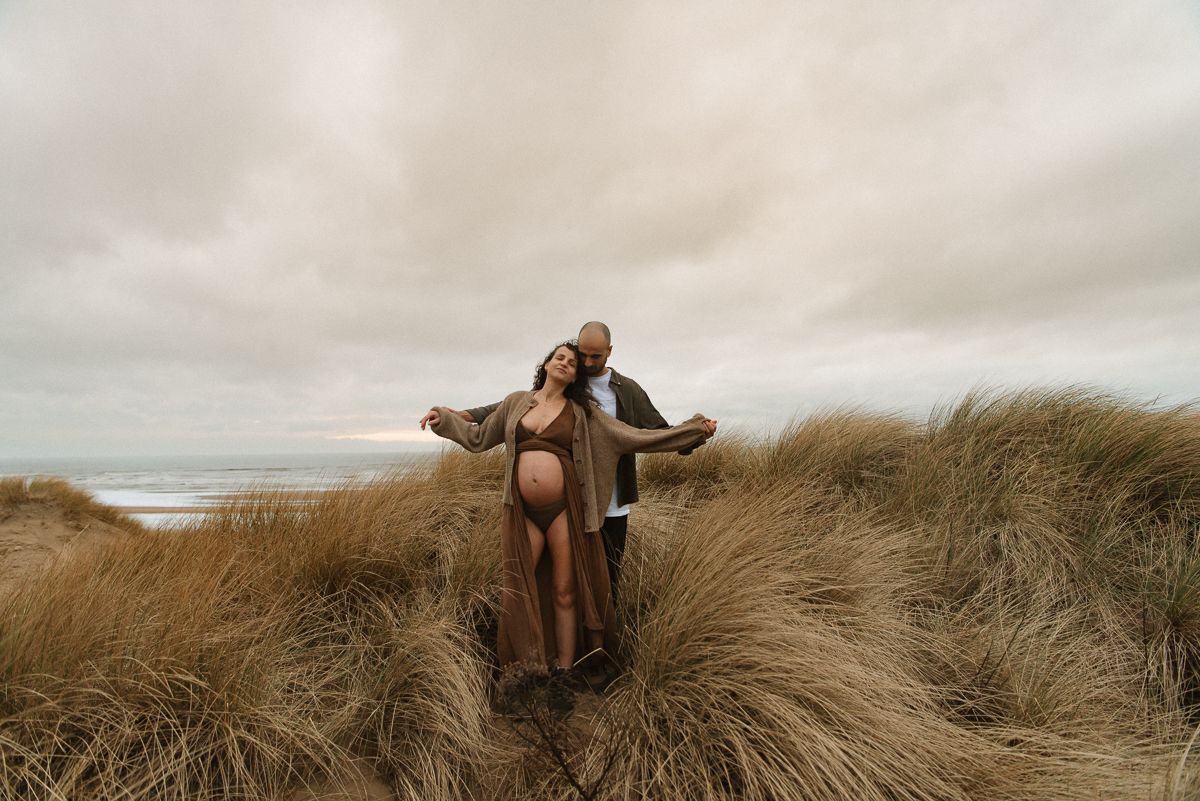 Figure stands with arms outstretched in beige coat among tall beach grass on cloudy coastal day.