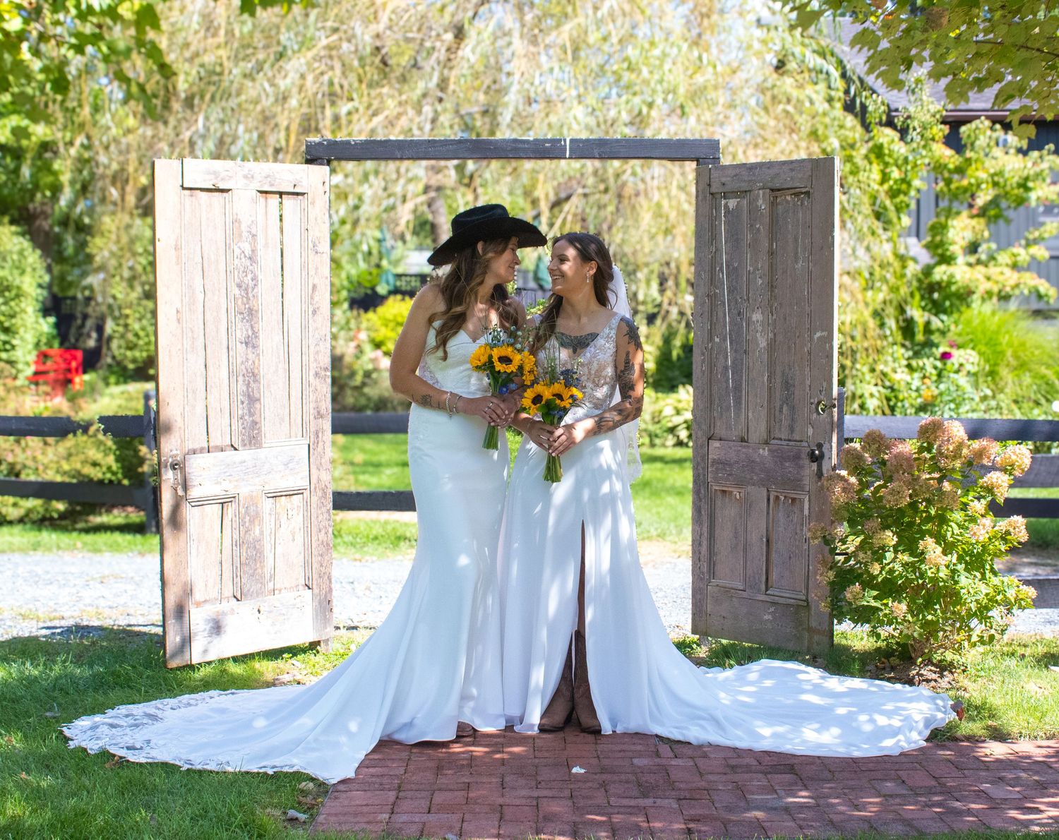Two people in wedding dresses standing in a garden archway holding bouquets.