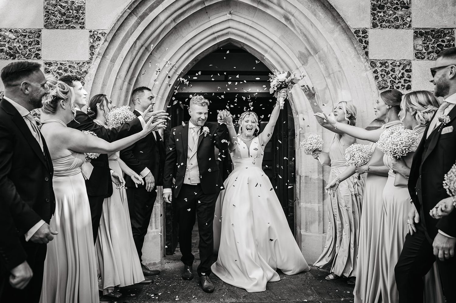 Wedding party throws confetti over newlyweds exiting stone church archway.
