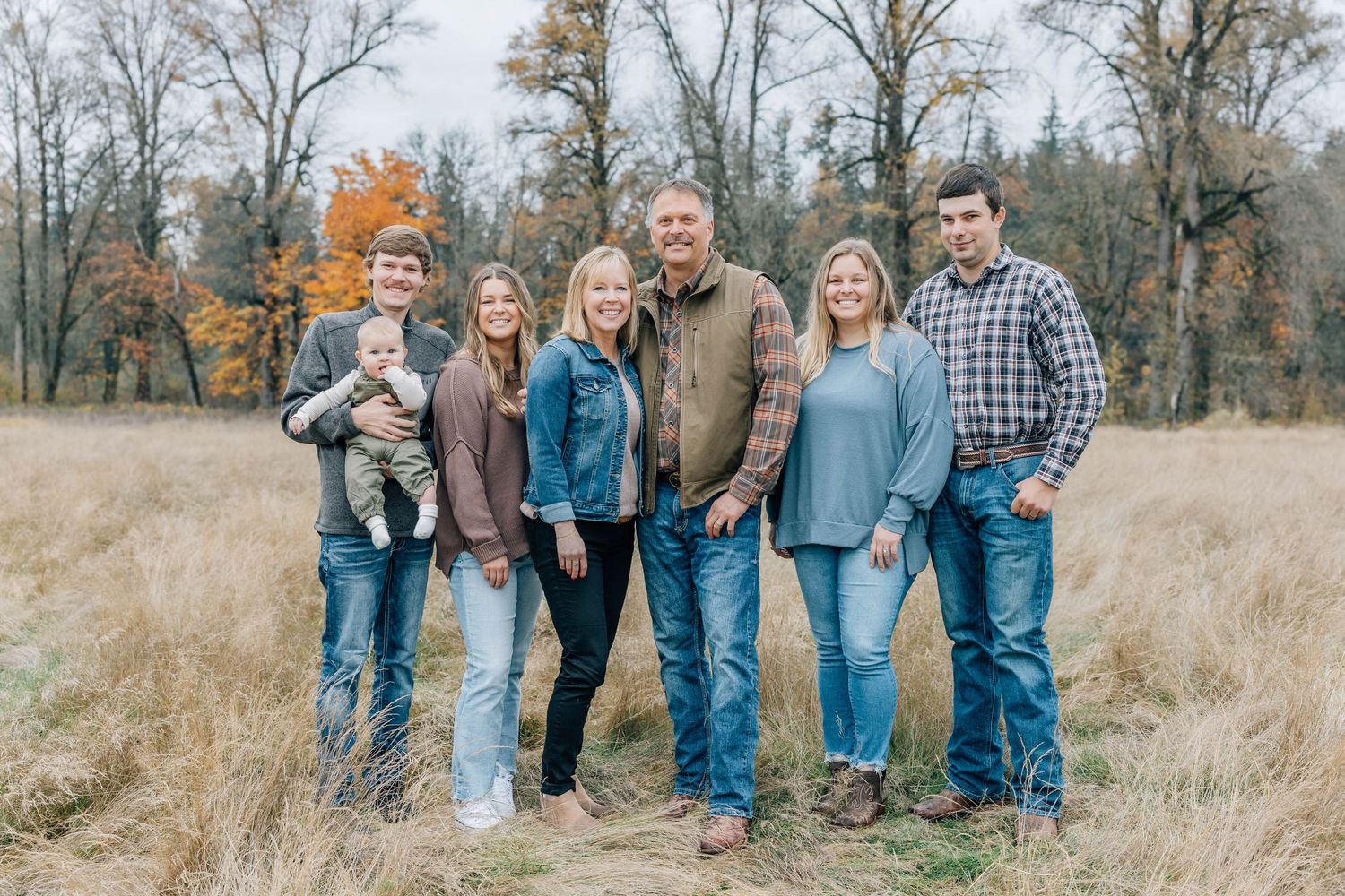 A family of seven poses together smiling in a golden autumn field wearing casual denim outfits.