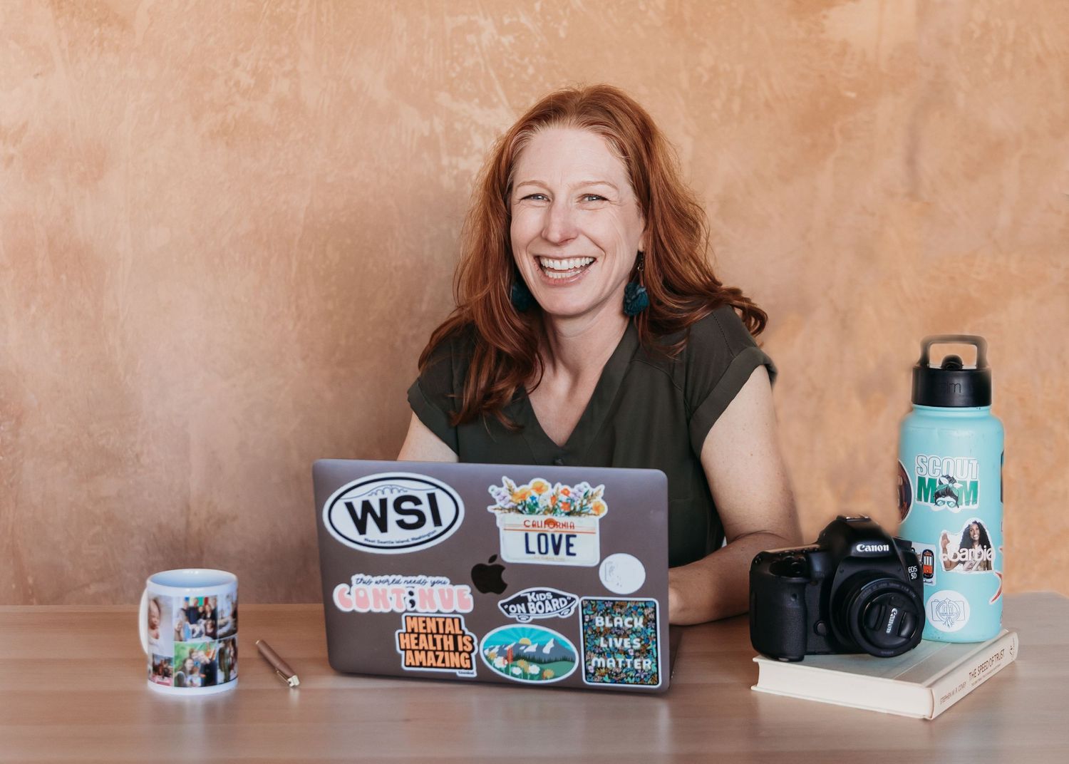 A laptop covered in stickers sits on a desk with a camera and water bottle against a textured brown wall.