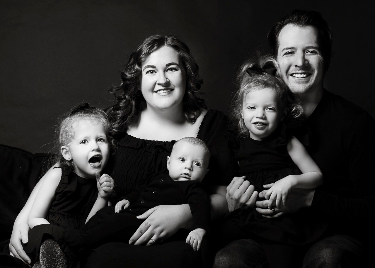 Black and white studio portrait of a family of five smiling and laughing together against a dark background.