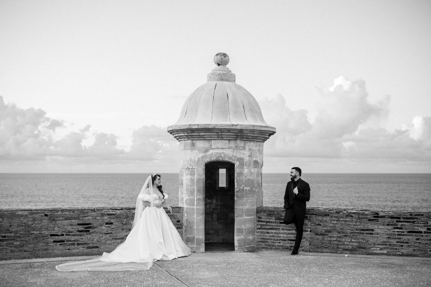 A black and white wedding photo by a historic stone guard tower overlooking the ocean.