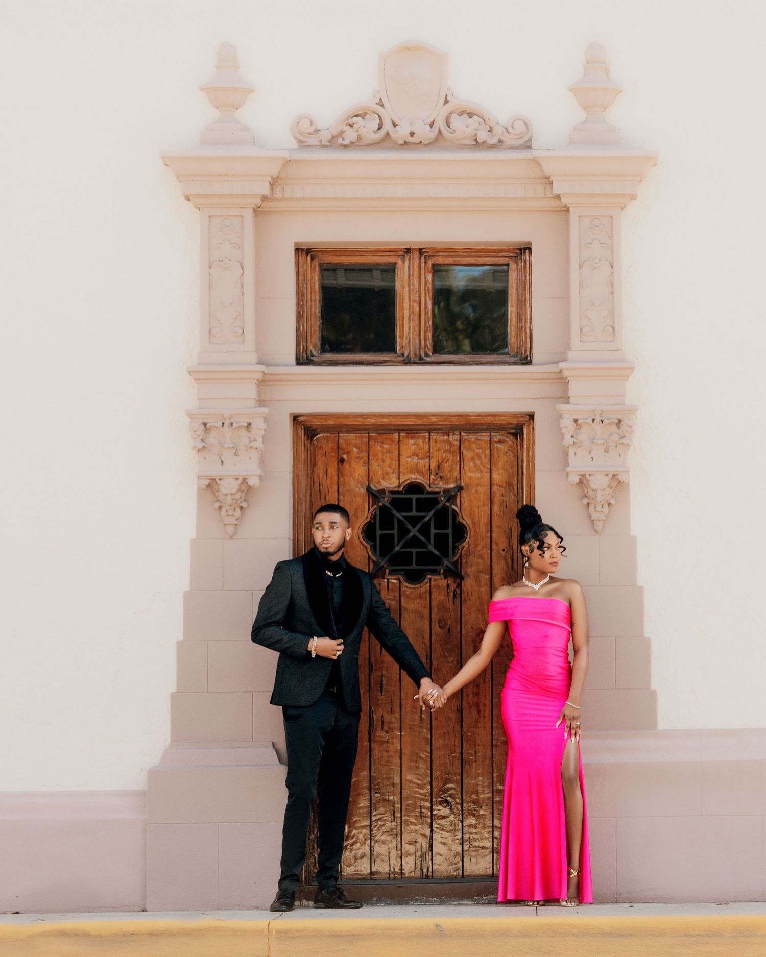 Formal couple posing by ornate doorway entrance in black suit and hot pink dress.