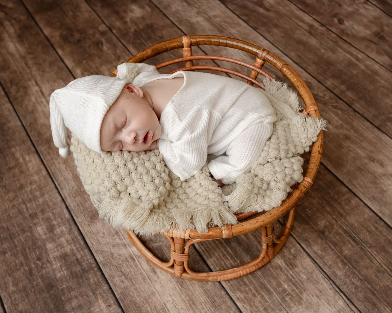 Sleeping newborn baby wrapped in white fabric lying in a round wicker basket on fluffy white bedding.