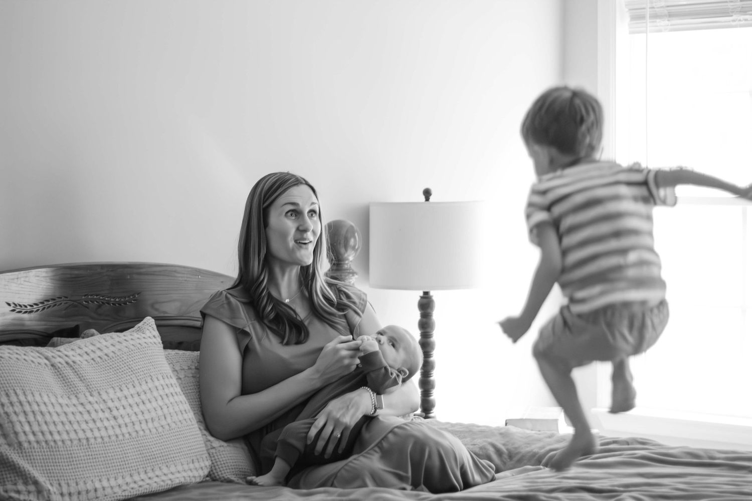Newborn Lifestyle: Mom and baby sitting on bed with toddler jumping on bed.