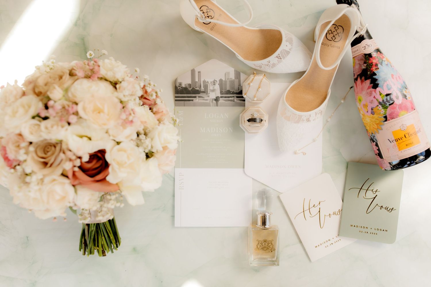 Wedding details flatlay with blush bouquet, white heels, perfume bottle, and stationery on marble surface.
