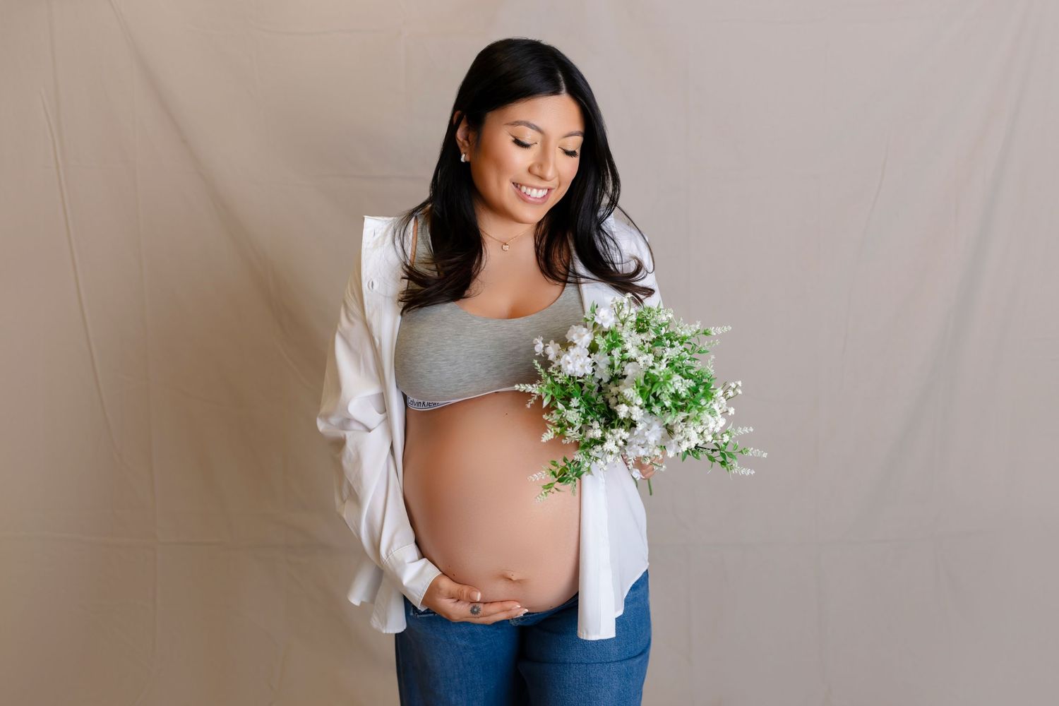 Expectant mother in casual attire holds delicate white flowers against neutral backdrop during maternity photoshoot.