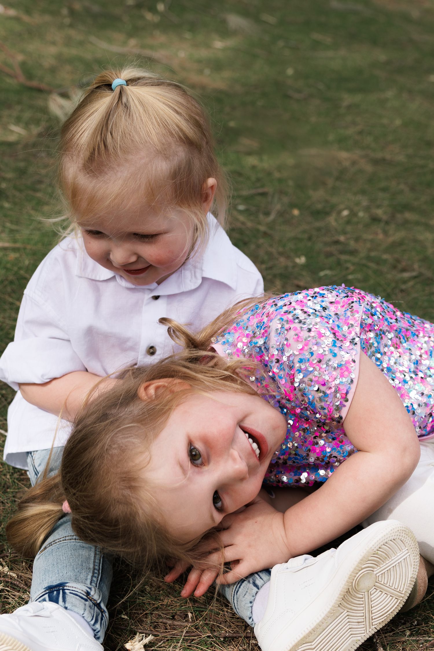 Two children playing and laughing together on grass, one wearing white shirt and other in floral print outfit.