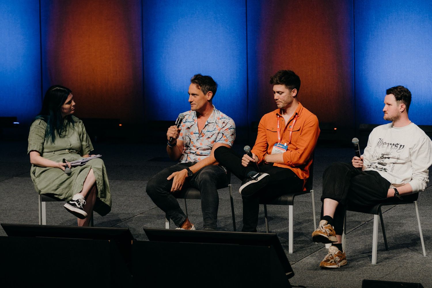 Four young people sit on chairs during a panel discussion on a stage with blue and orange lighting.