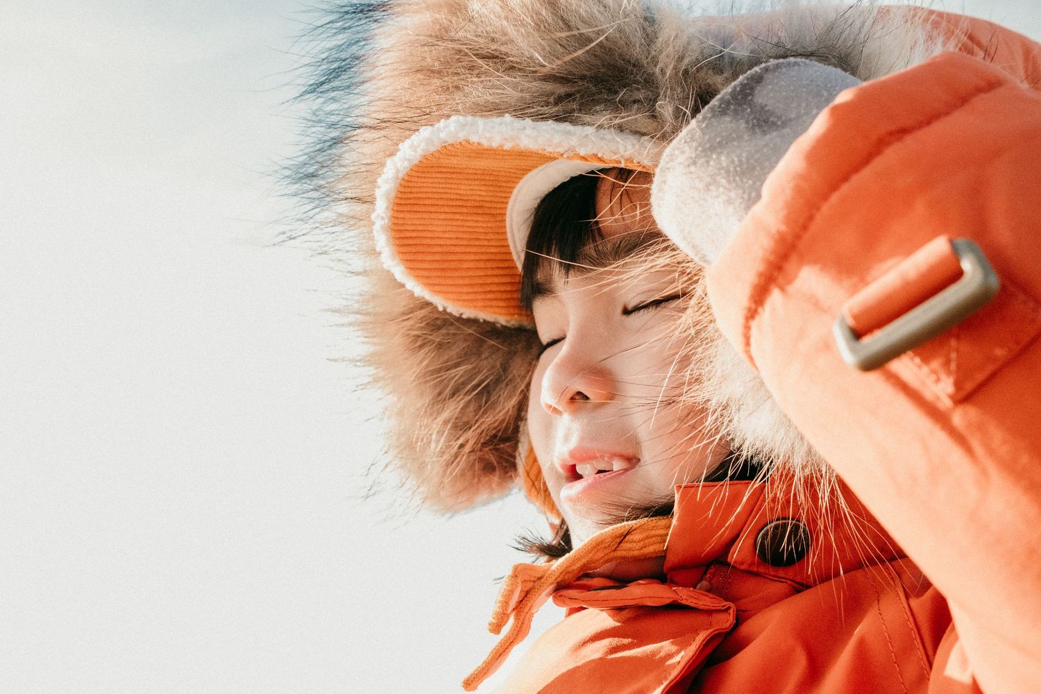 Young child in orange fur-trimmed winter jacket squinting in bright winter sunlight outdoors.