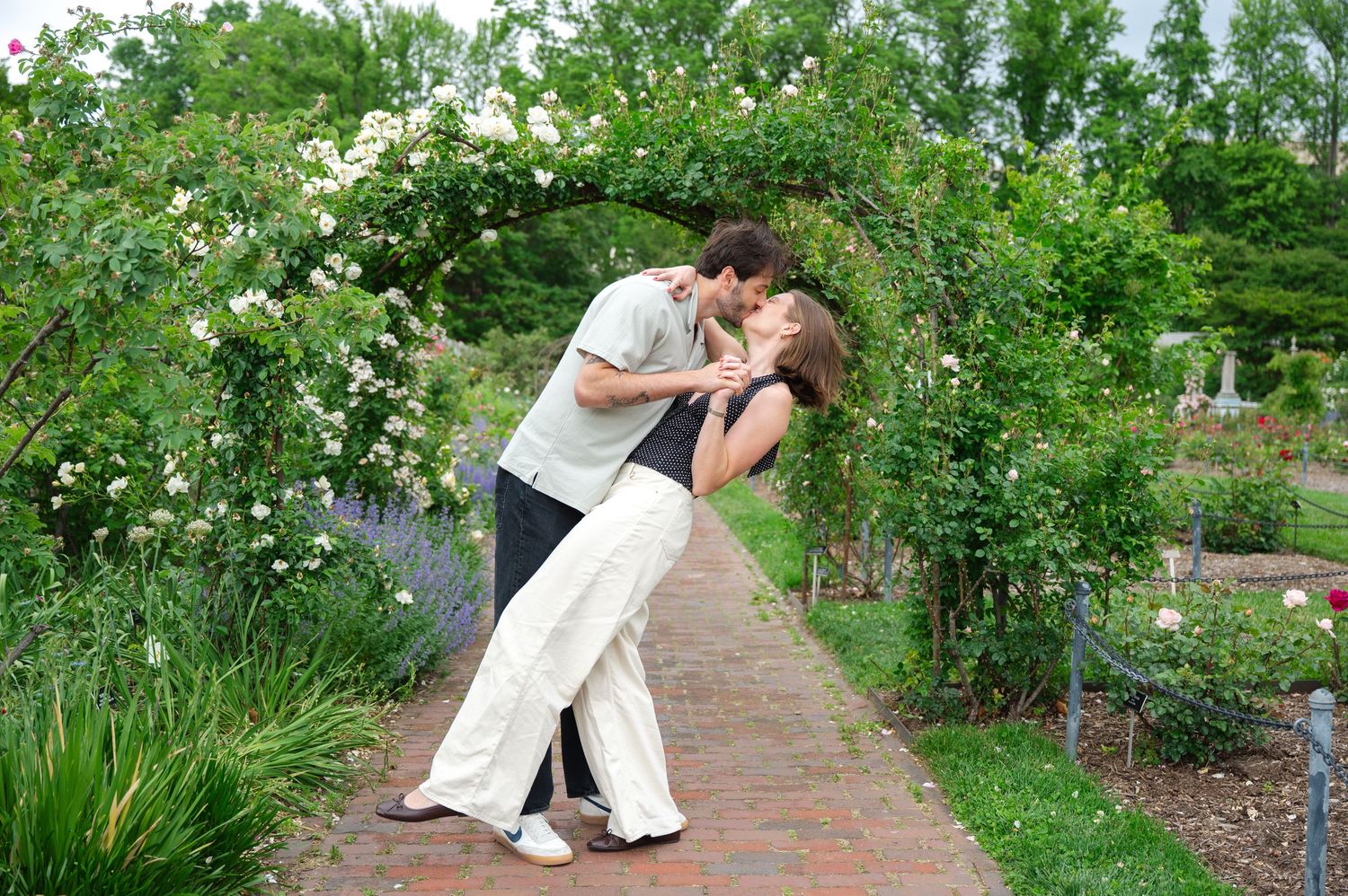 Couple sharing a romantic dip kiss under a rose garden arch on a brick pathway surrounded by lush greenery.