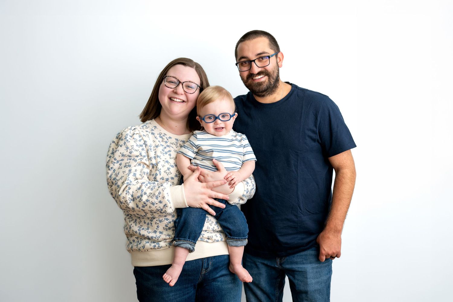 Happy young family with glasses posing together, mother holding their baby in a bright studio portrait.