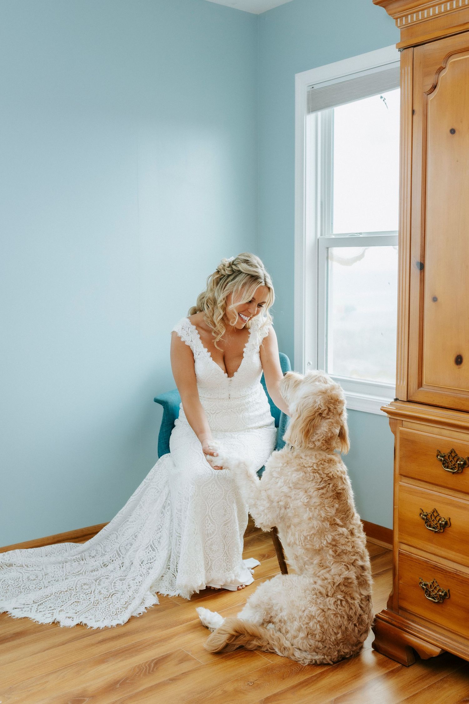 Bride in white lace gown sits smiling at her golden doodle dog beside a wooden armoire in a light blue room.