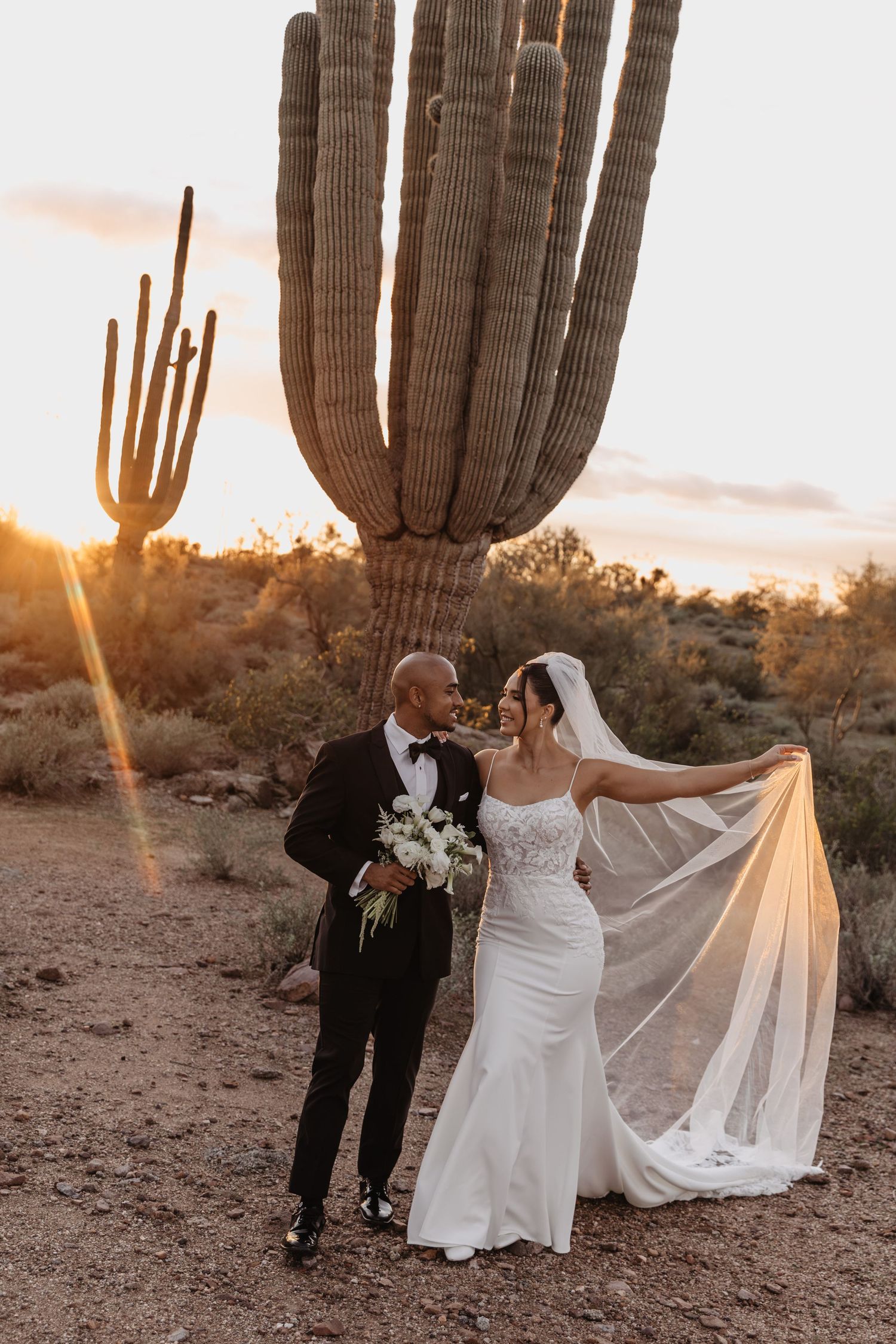 A romantic desert wedding photoshoot with saguaro cacti silhouetted against a glowing sunset sky in Arizona.