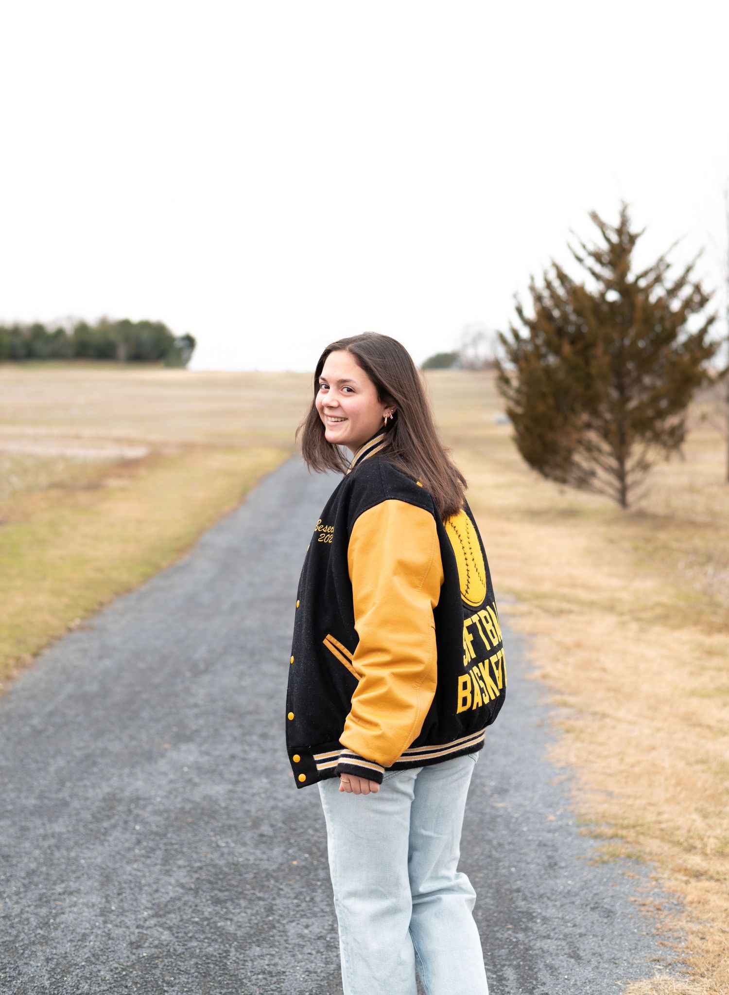 Person wearing a black and yellow varsity jacket with light jeans stands on a gravel path in a rural setting.