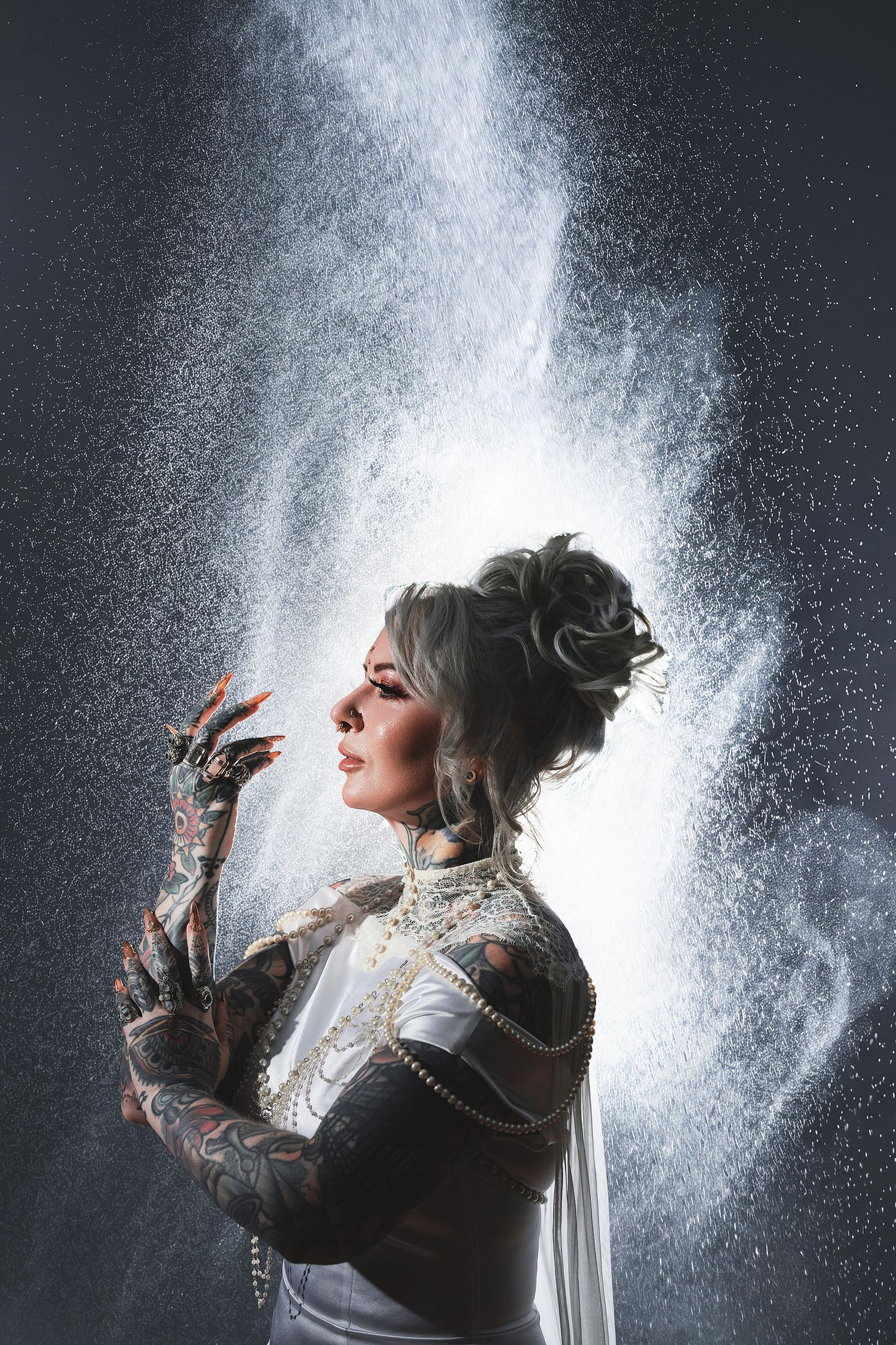 Tattooed woman in white gown with elegant updo poses dramatically as white powder cascades around her in studio.