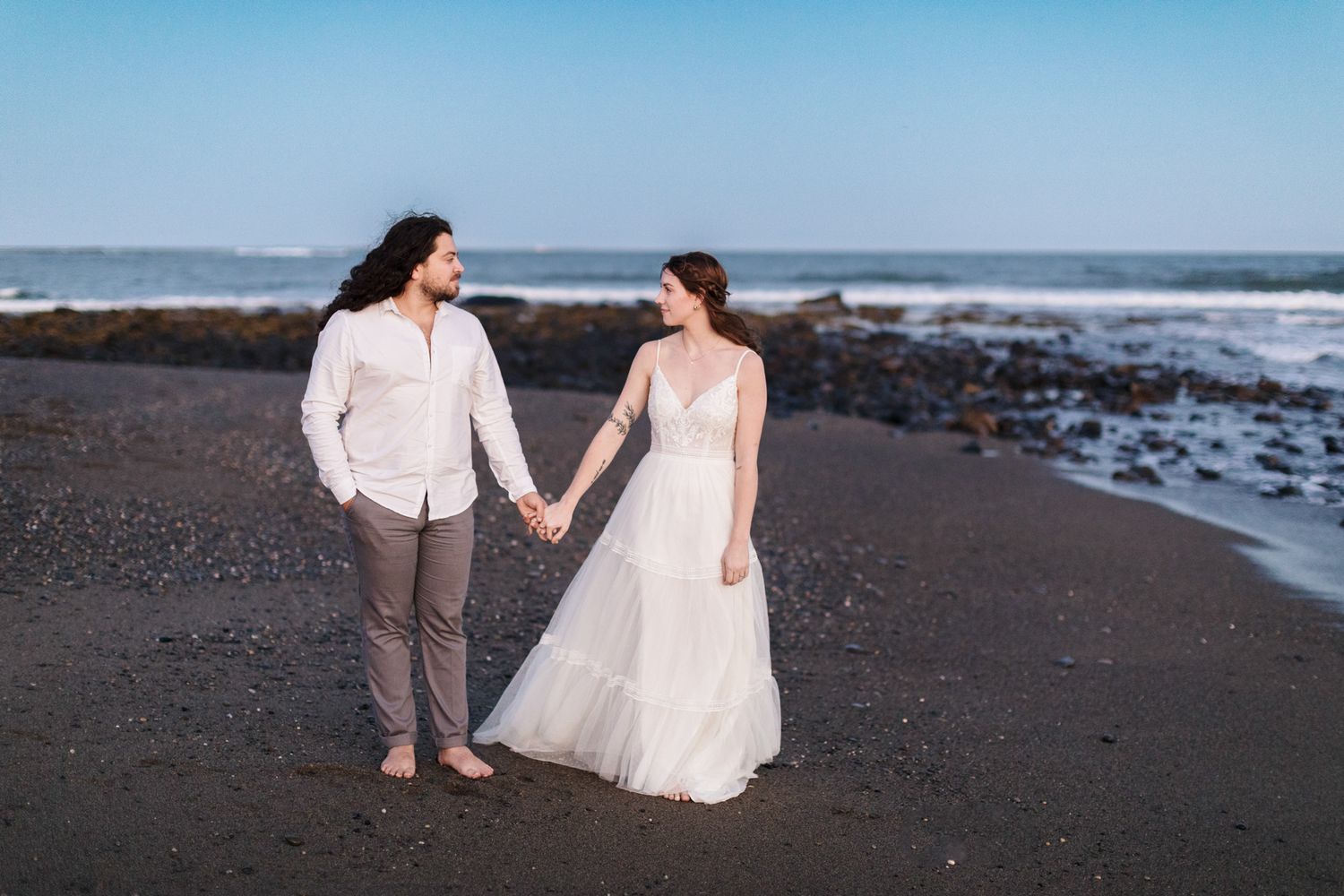 Woman in a bridal gown and man with white shirt and gray pants hold hands on a dark sand beach in Rye, New Hampshire at dusk.
