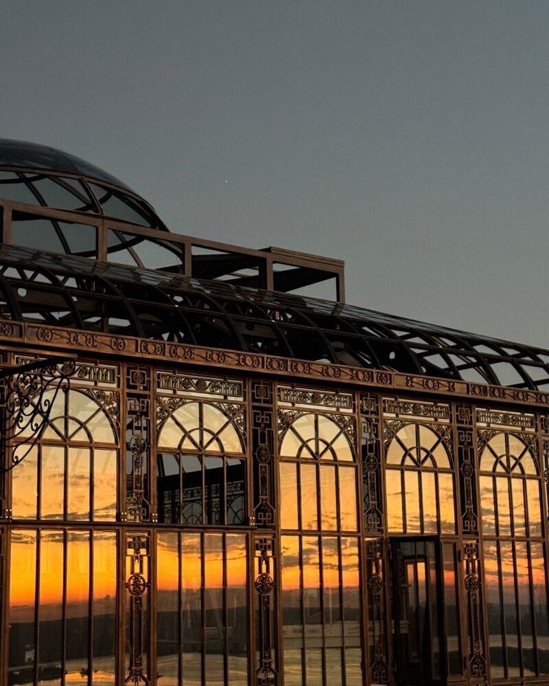 Ornate iron and glass architectural structure silhouetted against a vibrant orange sunset sky over the ocean.