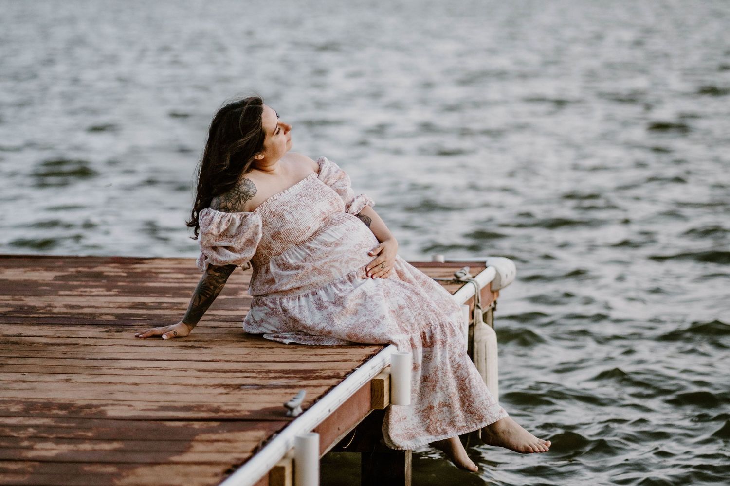 A pregnant woman in a flowing pink dress stands by a lake at dusk with wind blowing through her long dark hair.