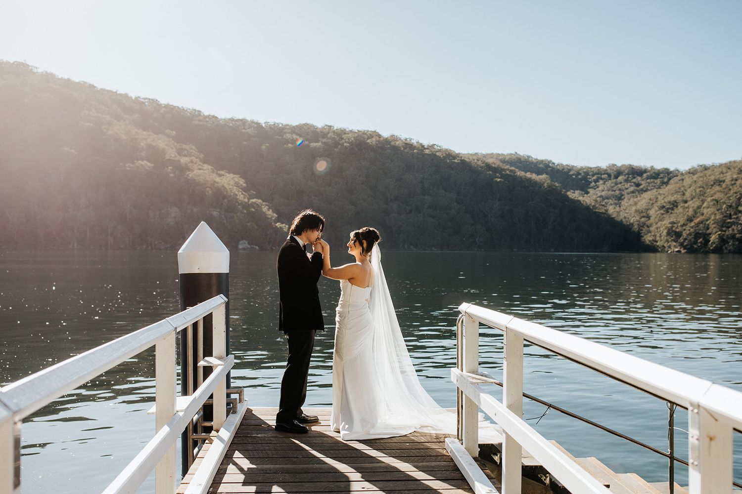 Bride and groom share a tender moment on a sunlit wooden dock overlooking a serene lake surrounded by lush green hills.