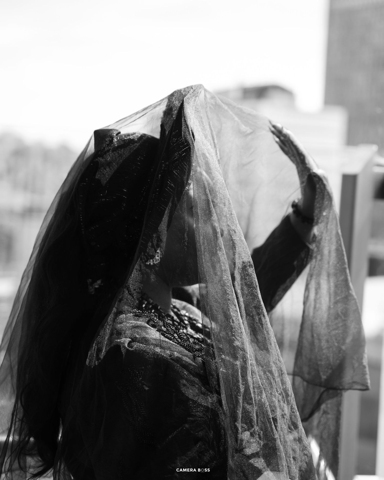 Black and white photo of a person draped in a sheer veil, raising their hand, wearing dark embroidered clothing.
